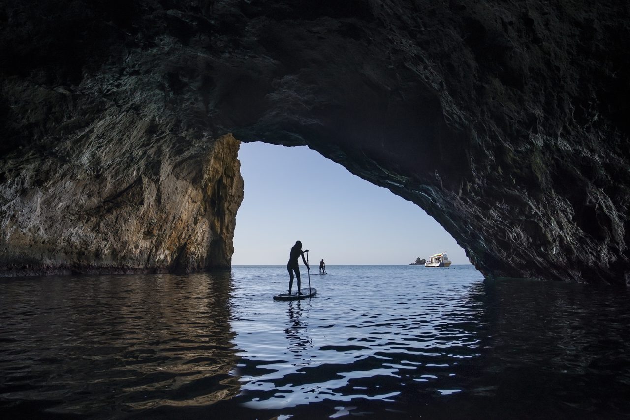 A person stand up paddle boarding in a dark cave with an opening to clear blue skys