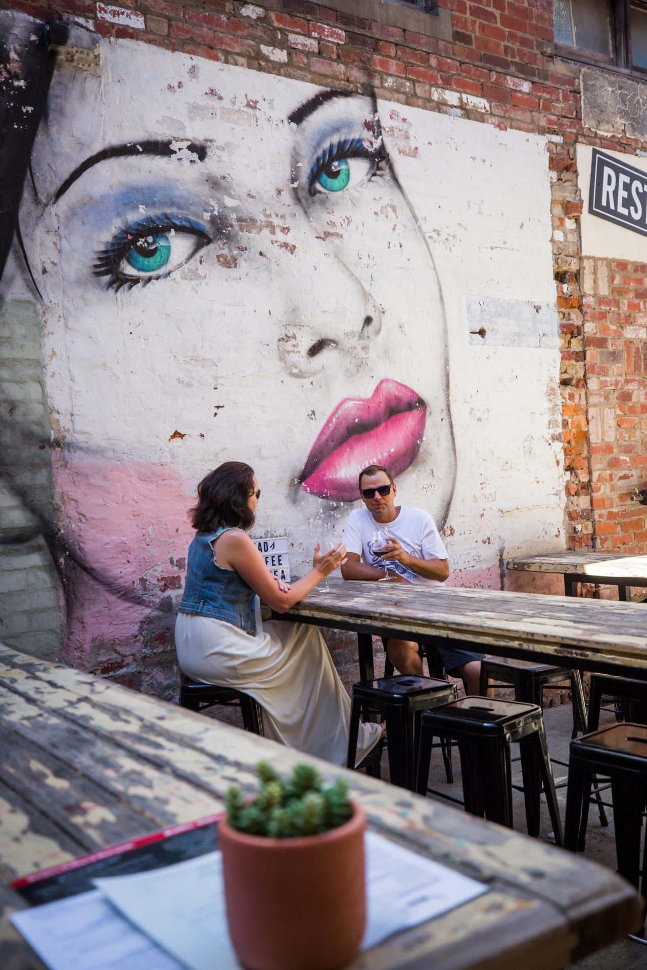 two people drinking wine on a wooden tableoutside in front of a mural of a woman's face.