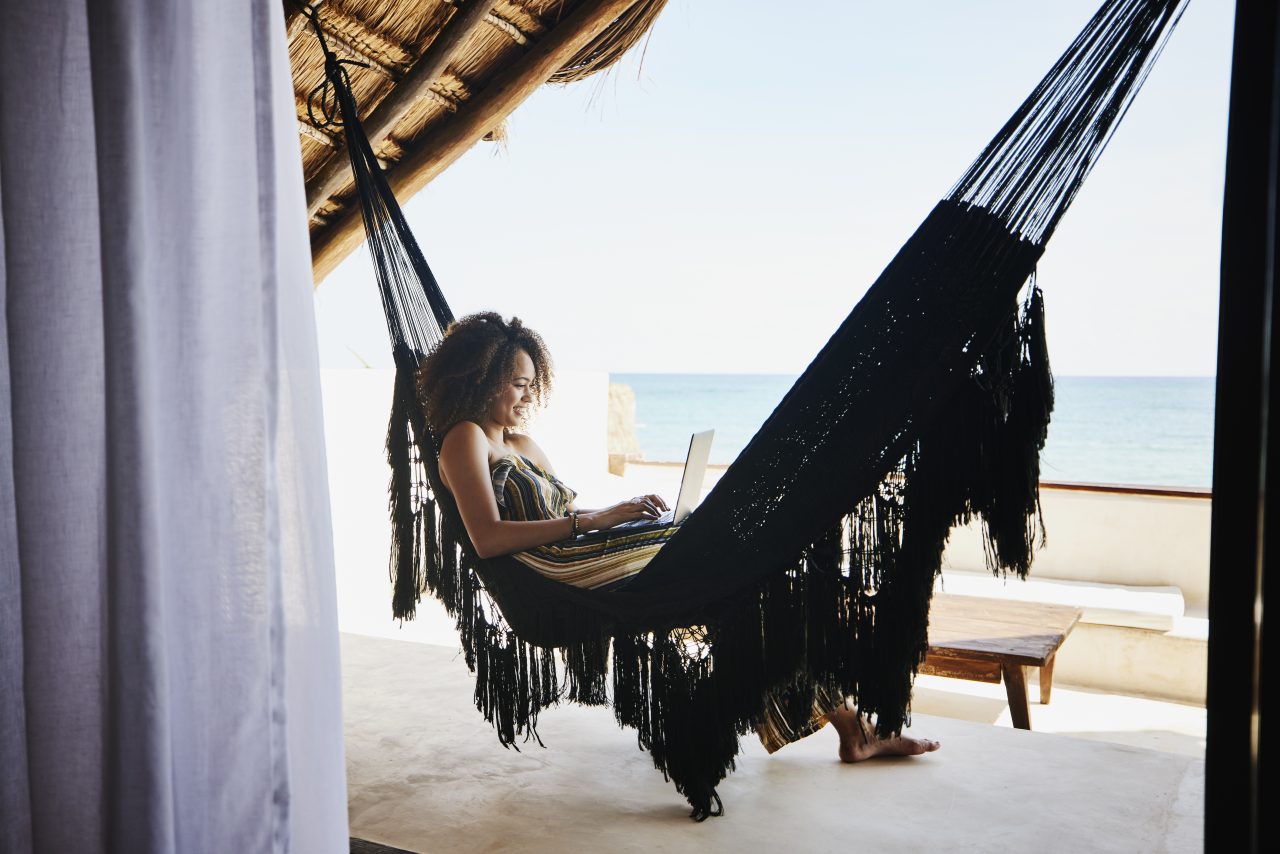 Wide shot of woman working on laptop while relaxing in hammock on deck of luxury tropical villa overlooking ocean