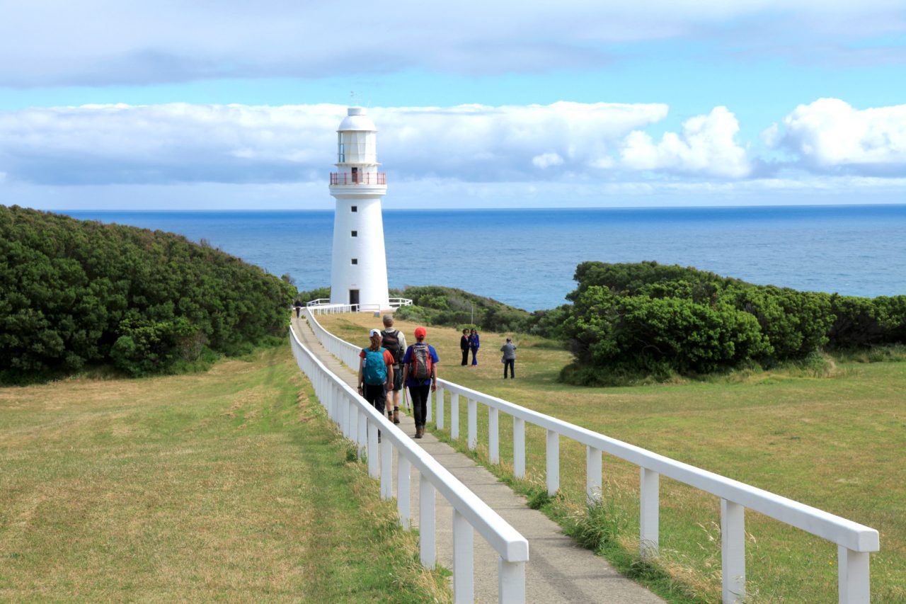 a group of people walking towards a lighthouse that is set on a cliff face that looks across the water.