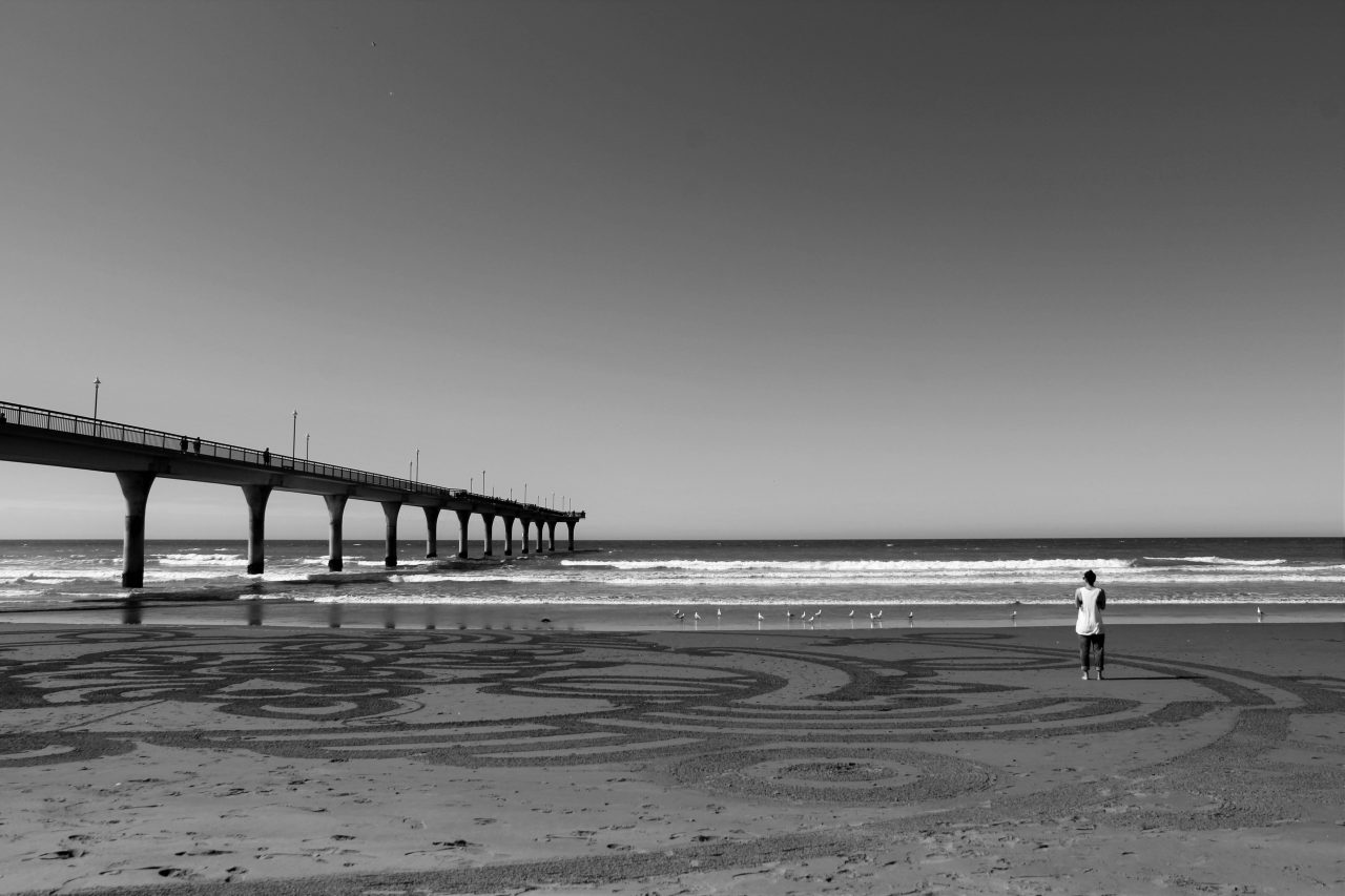 black and white image of a person looking out to the beach with a pier to the left of him.