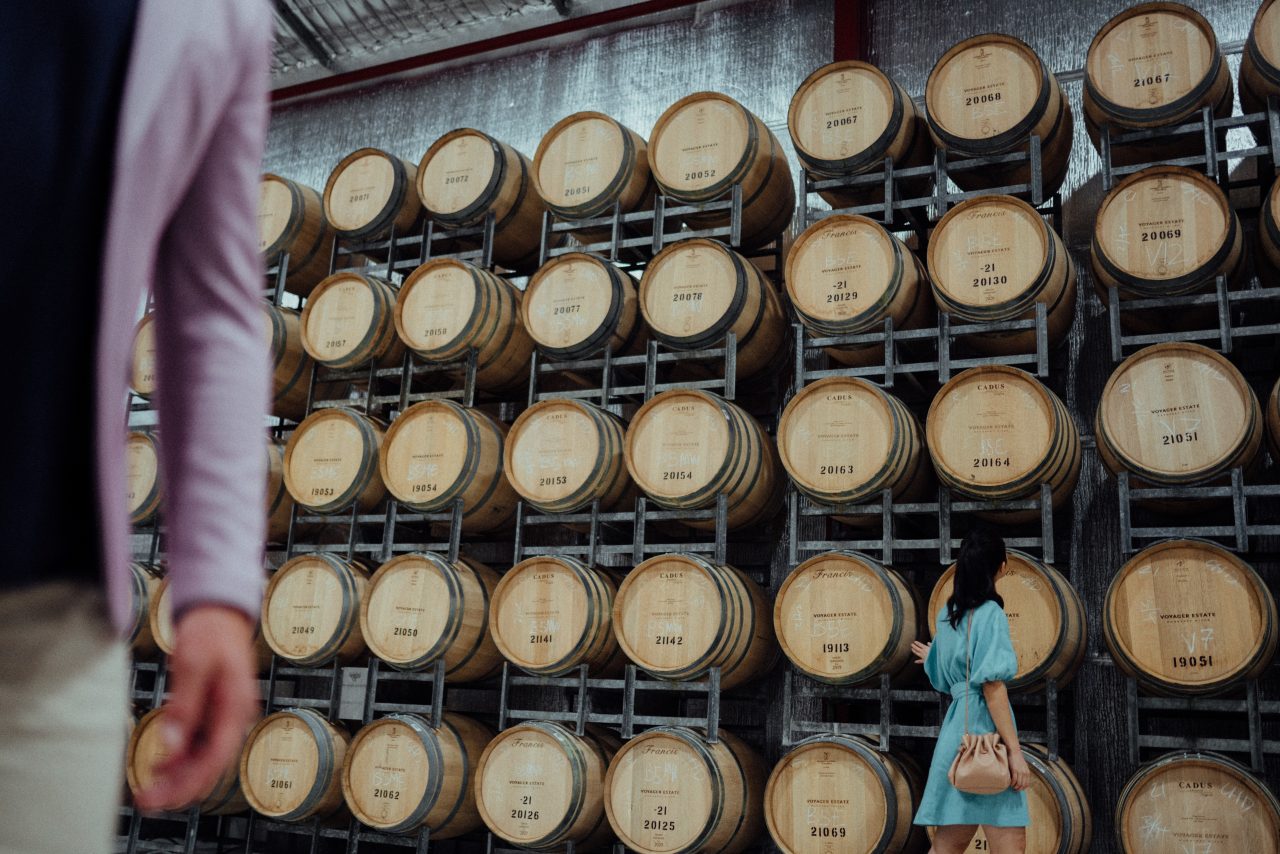rows and rows of barrels of wine stacked upon each other as a woman walks and looks at them,