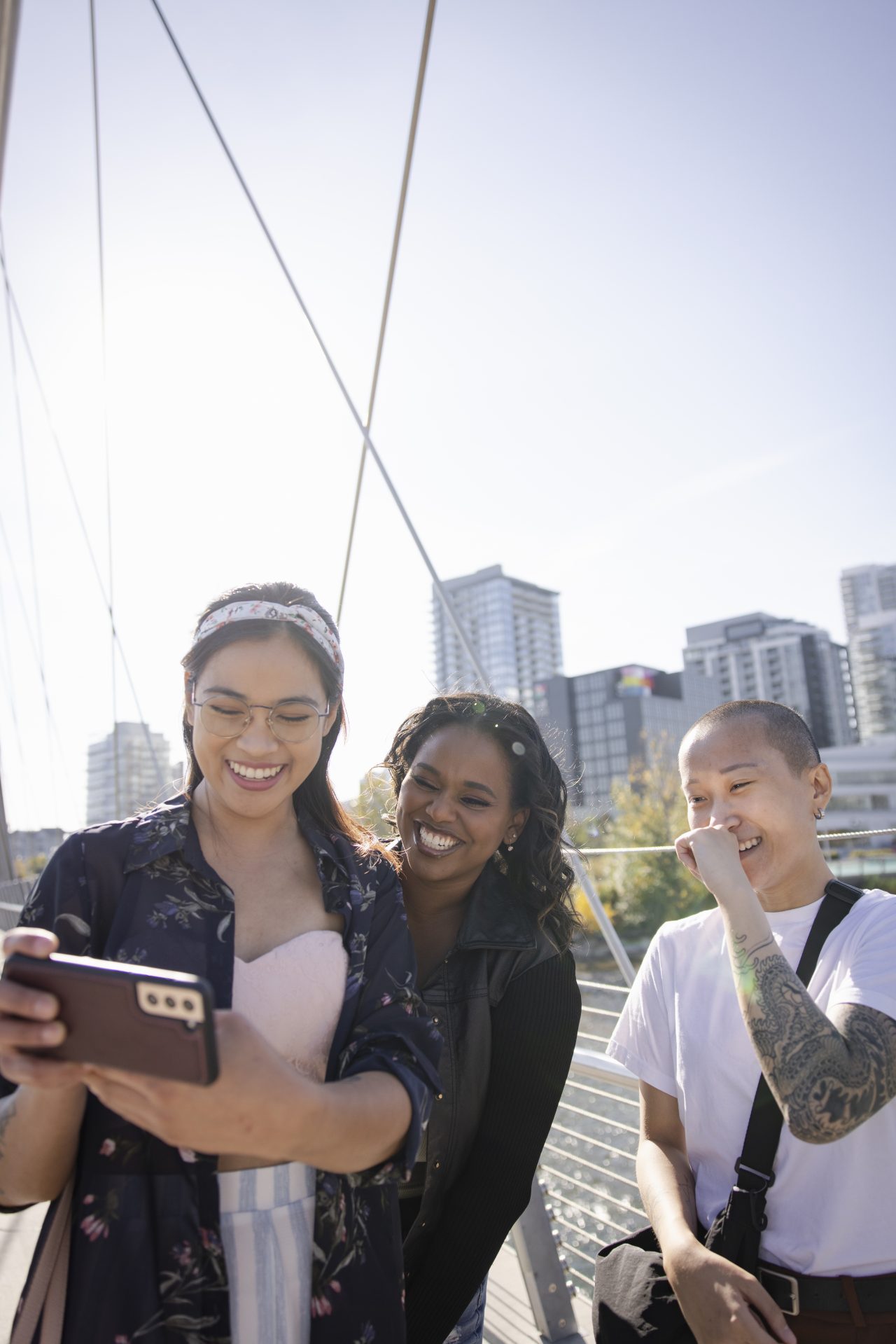 Happy young women friends laughing, taking selfie with smart phone on sunny urban footbridge