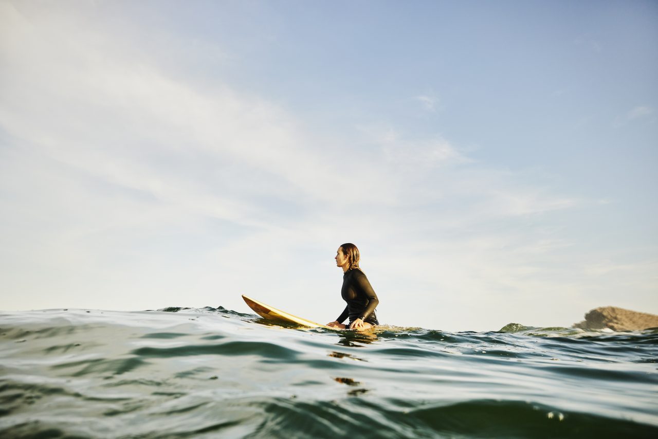 Wide shot of female surfer sitting on surfboard in ocean while waiting for wave during early morning surf session