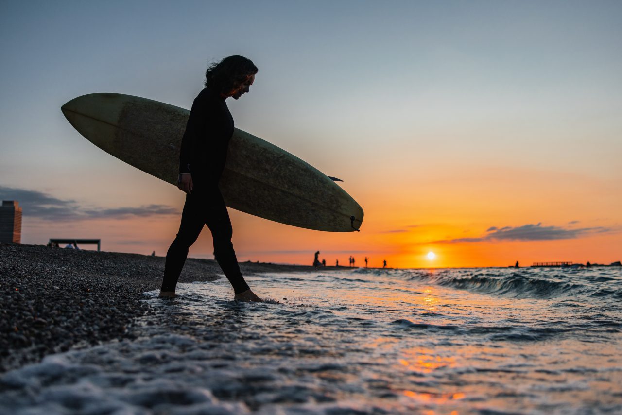 A black silhouette of a female surfer in a wetsuit carrying her surfboard into the water at the start of her early morning surfing session. She is about to enjoy a surfing session during a beautiful orange sunrise. Fun summertime activities for active people.
